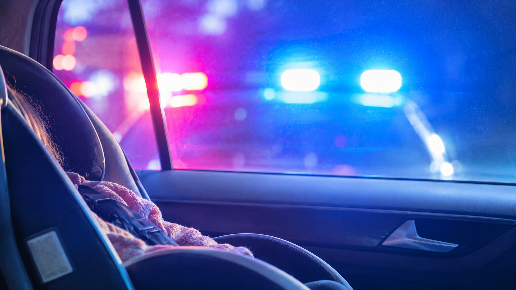 An empty child's car seat in a parked car at night, lit by red and blue police lights through the window
